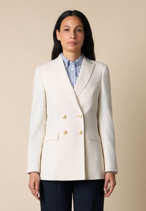 Kobieta ubrana w biały dwurzędowy blezer z złotymi guzikami, na niebiesko-białej pasiastce, stojąca na tle beżowego tła.