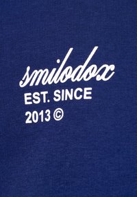 Weißer Text auf blauem Stoff lautet "smilodox EST. SINCE 2013 ©" in gemischter Schreibschrift und Blockbuchstaben.