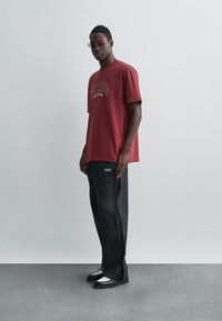 Crvena pamučna majica s otisnutim logom, uparena s crnim trapericama koje imaju bočne patentne zatvarače. Dodatci uključuju sunčane naočale i crnu obuću.