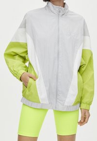Giacca leggera con zip di colore grigio, arricchita da accenti verdi e bianchi vivaci. Presenta un colletto alto, polsini elasticizzati e tasche laterali.