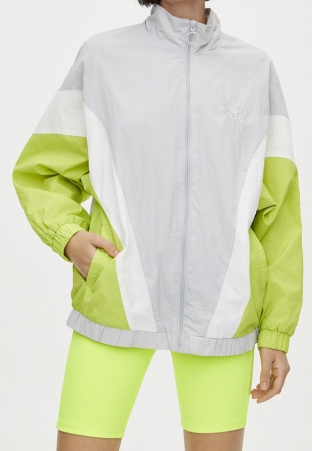 Giacca leggera con zip di colore grigio, arricchita da accenti verdi e bianchi vivaci. Presenta un colletto alto, polsini elasticizzati e tasche laterali.