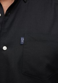 Black textured shirt with chest pocket, white buttons, and a small navy tag featuring a white deer head logo.