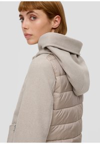 Jeune femme aux cheveux courts portant un gilet matelassé beige et un sweat à capuche assorti, regardant par-dessus son épaule sur un fond gris clair.