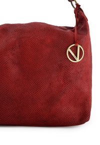 Rød tekstureret lædertaske med en glat overflade, prydet med et gyldent, cirkulært hardware-element og en dekorativ rød båndvedhæng.