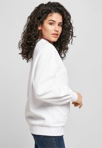 Balta sportinė palaidinė su ilgomis pūstomis rankovėmis, ribnutėmis apyrankėmis ir apačia, pagaminta iš minkštos medžiagos, dėvima su mėlynais džinsais. Paprastas dizainas, be matomų logotipų.