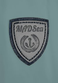 Shield-shaped patch made of embossed gray leather, featuring "MADSea" text and a design with an anchor and circular motifs, set against a teal background.