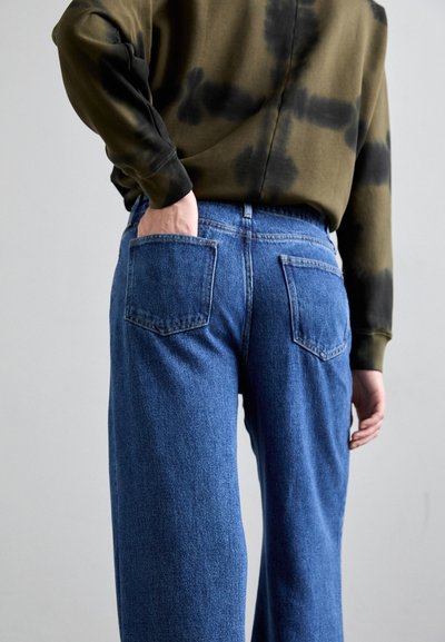 Blugi din denim albastru cu o croială largă, având două buzunare pe spate și cusături în contrast. Asortați cu un hanorac tie-dye de culoare verde măslin.