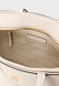 Intérieur d'un sac à main en cuir beige avec une fermeture éclair dorée, doublure en tissu avec le nom de la marque en relief, et un accent noir sur le bord supérieur.