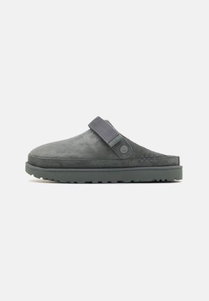 Zapato slip-on de ante gris con un perfil bajo y suela texturizada. Presenta una correa de tela y un detalle del logo en el costado.