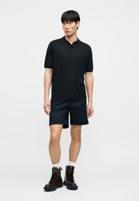 Άνδρας με μαύρο κοντομάνικο πόλο, ναυτικό σορτς, λευκές κάλτσες και σκούρα καφέ μπότες με κορδόνια, στέκεται σε μονόχρωμο φόντο.