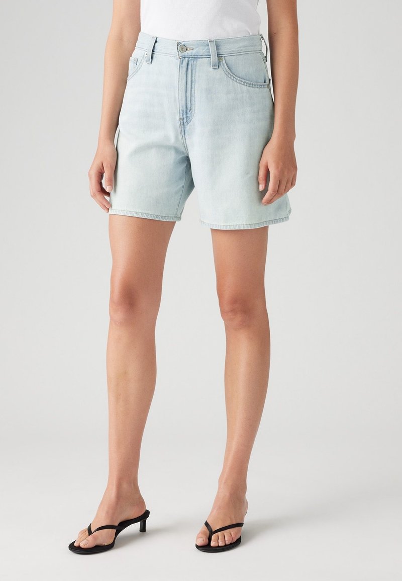 Personne portant un short en jean bleu clair, un haut blanc et des sandales à talons noires, debout devant un fond gris uni.