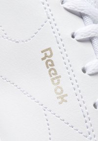 Baskets synthétiques blanches avec une tige texturée, des accents cousus et un logo "Reebok" embossé en or sur le côté.