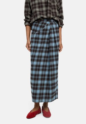 Osoba nosząca długą niebiesko-czarną kratowaną spódnicę, wpuszczoną brązową kratowaną koszulę i czerwone buty na wsuwane, stojąca na jednolitym tle.