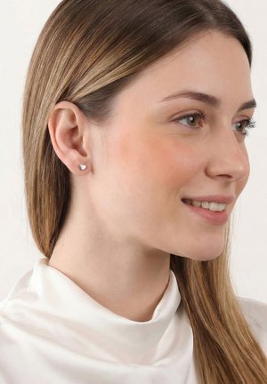 Jeune femme aux longs cheveux châtain clair portant de petites boucles d’oreilles en forme de cœur en argent et un chemisier blanc à col montant, souriant légèrement.
