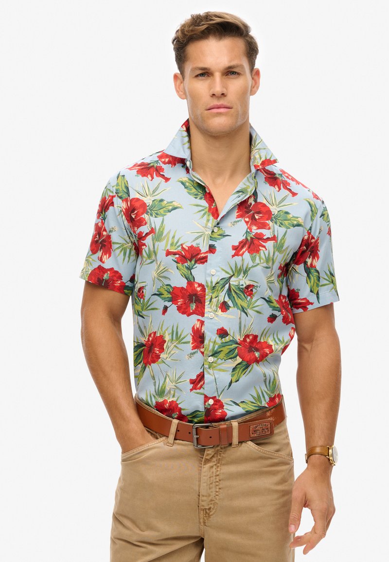 Camisa de manga corta con fondo azul claro, adornada con flores de hibisco rojas y hojas verdes. Diseño con botones en la parte delantera, con cuello.