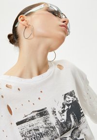 Hvid bomulds t-shirt med slidte huller, prydet med et sort-hvidt grafisk print. Modellen bærer oversized solbriller og sølvfarvede hoop øreringe.