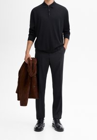 Schwarzes Langarm-Polohemd, maßgeschneiderte schwarze Hose und eine braune Wildlederjacke, die über einen Arm gehängt ist; schwarze Schuhe runden das Outfit ab.