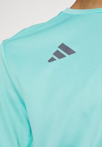 Světle tyrkysové sportovní tričko z prodyšného materiálu. Na zadní straně má šedé logo s texturovaným povrchem a detailem švu na rameni.