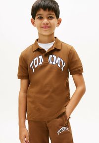 Brun polo skjorte med hvid "TOMMY" skrift, korte ærmer og krave. Bæres med matchende brune bukser. Glat tekstur.