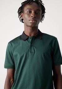 Mladý muž s krátkymi dredmi, ktorý má na sebe tmavozelené polo tričko s čiernym golierom, stojí proti jednému bielému pozadiu.