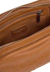 Interior de bolsa de cuero marrón claro con un bolsillo con cremallera y una etiqueta de parche. Presenta una textura suave y costuras en contraste. Hardware plateado visible.