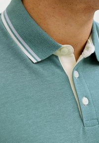 Κοντινό πλάνο ατόμου που φοράει πετρόλ πόλο πουκάμισο με λευκή λωρίδα στο γιακά και τρία λευκά κουμπιά, δείχνοντας τον λαιμό και το πάνω μέρος του στήθους.