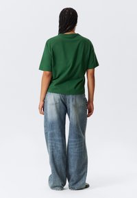 T-shirt verde in cotone con una vestibilità comoda, abbinata a jeans in denim a gamba larga di colore blu chiaro, con macchie sbiadite e dettagli di cucitura visibili.