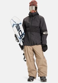 Snøbrett med svart og gråmønstret jakke, beige bukser, hansker og strikket lue. Reflekterende goggles dekker øynene. Snøbretter har en hvit bunn.