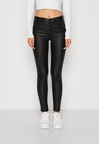 Zwarte strakke jeans gemaakt van een stretchy, faux leren materiaal. Kenmerken zijn een sluiting met een knoop aan de voorkant en riemlussen, gecombineerd met witte sneakers.