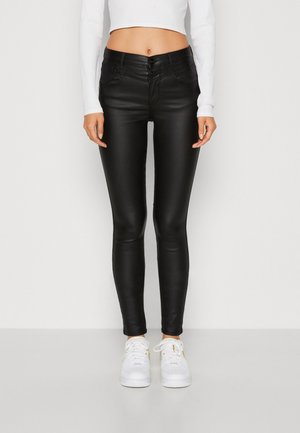 Jean skinny noir en similicuir extensible. Fermeture à bouton à l'avant et passants de ceinture, associés à des baskets blanches.