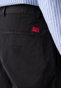Black cotton pants with a slim fit, featuring a red "HUGO" tag on the back pocket and a subtle texture. Accented with a folded waistband.