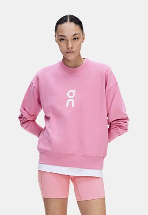 Femme aux cheveux foncés en chignon, portant un sweat-shirt rose avec un logo blanc et un cuissard rose assorti, debout les mains dans le dos.