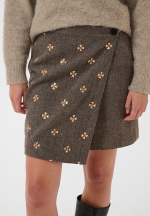 Personne portant un pull beige et une jupe portefeuille marron ornée de motifs floraux en sequins dorés, debout les bras détendus sur les côtés.