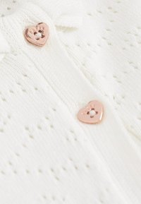 Bela pletena cardigan s odprtim vzorcem, ki ima dva gumbka v obliki srca iz rožnatega zlata in majhen lok na ovratniku.
