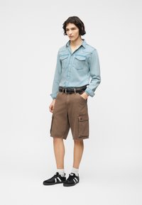 Νεαρός άνδρας που φοράει ανοιχτό μπλε τζιν πουκάμισο, καφέ cargo σορτς, μαύρη ζώνη, άσπρες κάλτσες και μαύρα αθλητικά παπούτσια με λευκές ρίγες, στέκεται με το χέρι στην τσέπη.