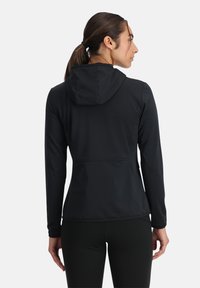 Donna con i capelli castani raccolti in una coda di cavallo, indossa una giacca nera con cappuccio e leggings neri, di spalle contro uno sfondo bianco semplice.
