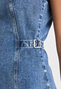 Gros plan d'une robe en denim bleu avec une ceinture à boucle argentée cintrant la taille et des coutures visibles sur le tissu.