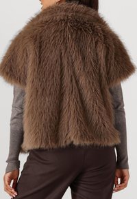 Bruine nepbontcape met korte mouwen, textuur, gelaagd ontwerp. Gemaakt over een getailleerd grijs lang shirt en bruine broeken.