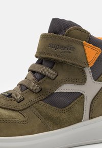 Høye sneakers med olivengrønn semsket skinn og mesh-paneler. Funksjoner inkluderer beige aksenter, en borrelåsrem og teksturerte snører.