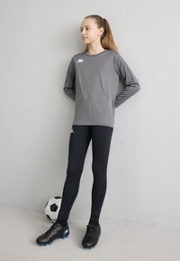 Siva športna majica z dolgimi rokavi, črne legice, črni nogometni kopački z modrimi poudarki. Stoji ob črno-beli nogometni žogi.