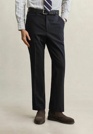 Uomo che indossa pantaloni formali neri, scarpe in camoscio marroni, camicia a righe e cravatta a righe, in piedi con una mano in tasca contro uno sfondo semplice.