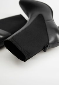 Bottes noires à cheville avec un bout en cuir élégant et une tige en tricot texturé. Le design comprend une petite languette de tirage au talon.