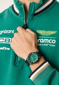 Sudadera verde con media cremallera, con un cuello y logotipo en blanco y negro. Reloj negro y verde azulado con una esfera verde texturizada y correa de goma.