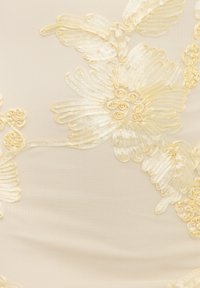 Broderie florale couleur crème avec ruban et petits détails en spirale sur fond de tissu beige transparent.