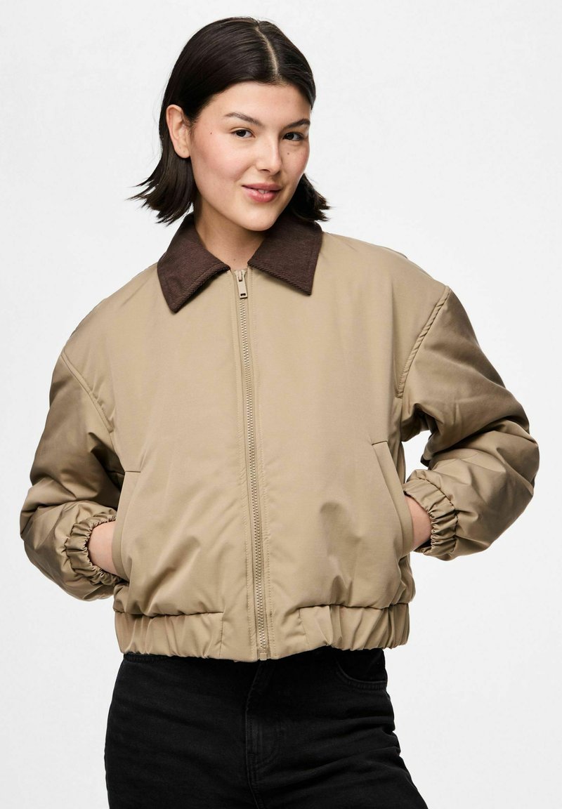 Sieviete ar tumšiem matiem, tērpusies bīgā pieguļošā jakā ar brūnu apkakli un melnām biksēm, rokas ieliktas kabatās, pret vienkāršu baltu fonu.