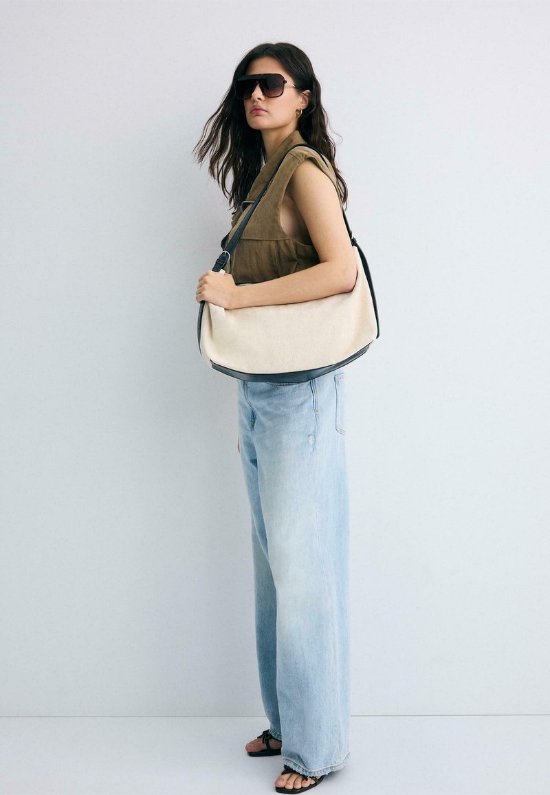 Femme portant de grandes lunettes de soleil, un top marron sans manches, un jean large bleu clair, des sandales noires et un grand sac beige porté à l'épaule.
