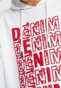 Bílá mikina s kapucí a velkým, opotřebovaným červeným potiskem s nápisem "DENIM" opakovaně. Má stahovací šňůrky a měkkou, strukturovanou látku.