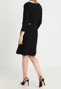 Svart klänning med långa ärmar, som visar en figursydd liv och utsvängt fåll, accentuerad med ett tunt midjebälte. Slätt tyg med en minimalistisk design.