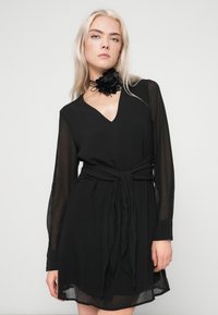 Γυναίκα με μακριά ξανθά μαλλιά, φορώντας ένα διάφανο μαύρο φόρεμα με μακριά μανίκια, δεμένο στη μέση και μια μαύρη λουλουδένια αλυσίδα γύρω από τον λαιμό της.