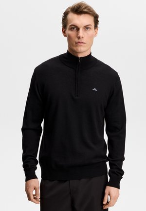 Młody mężczyzna w czarnym, długim rękawie z krótkim zamkiem błyskawicznym z przodu i małym logo na piersi, stojący na białym tle.